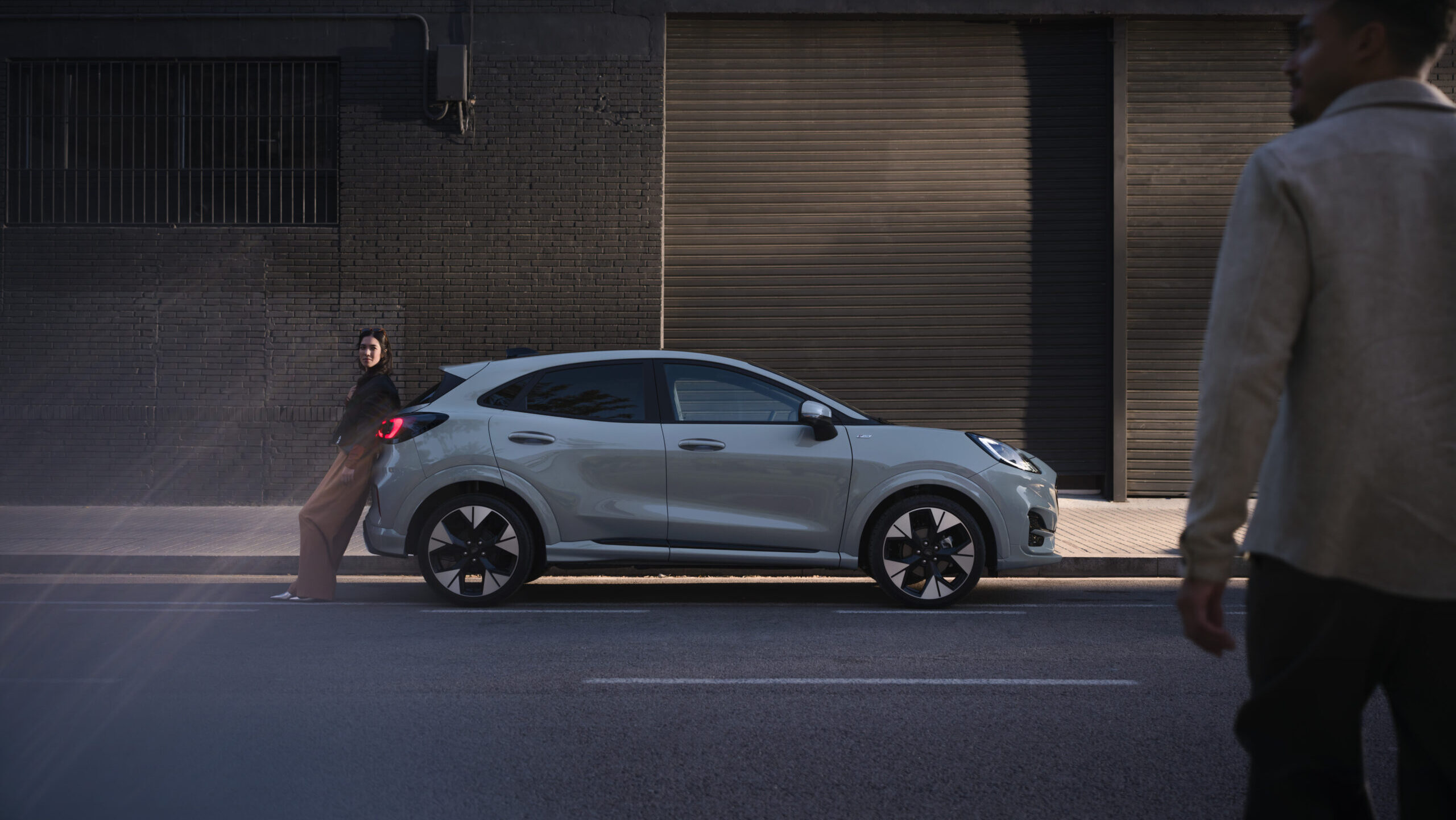 Seitliche Ansicht eines hellen SUVs in der Stadt, Frau lehnt am Fahrzeug, Passant im Vordergrund